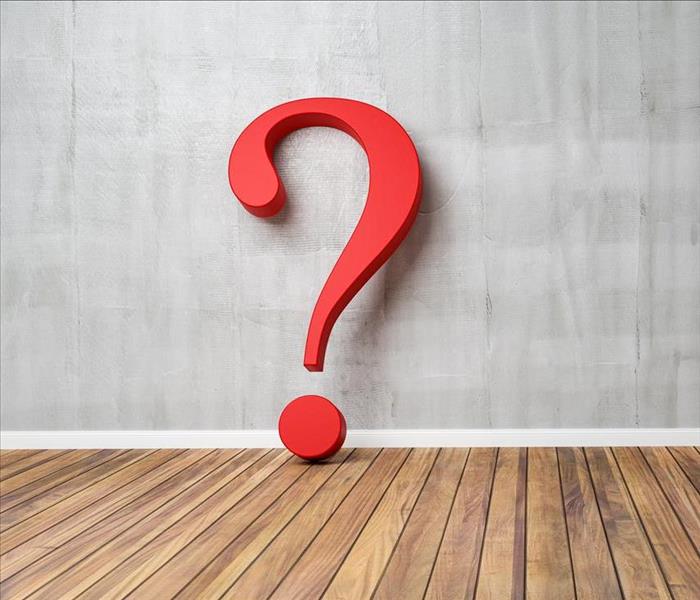 A large question mark symbol made of plastic or wood is in a room leaning against a gray wall and sitting on a wood floor
